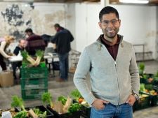 A man stands proudly in front of boxes of vegetables at a community food hub. There are other volunteers blurred in the background having an important conversation about food security. 